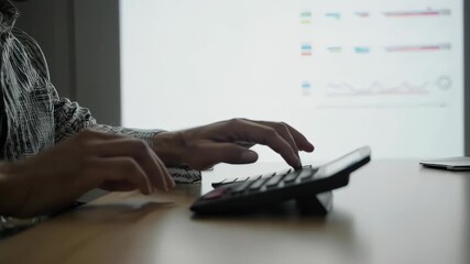 A person using a calculator with charts displayed in the background on a projector screen in a room - Powered by Adobe