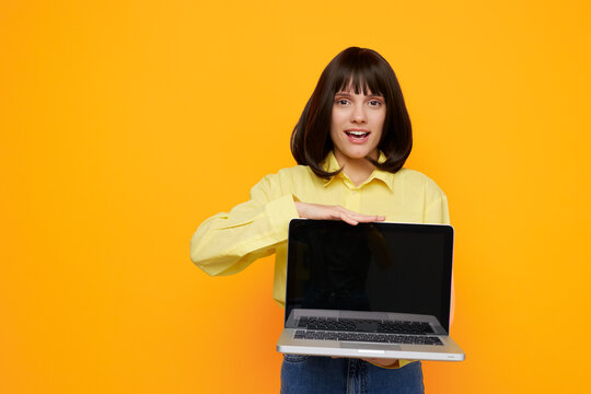 Cheerful young woman holds a laptop, presenting a bright digital scene. She smiles, wearing a yellow shirt, standing against a bold orange background.