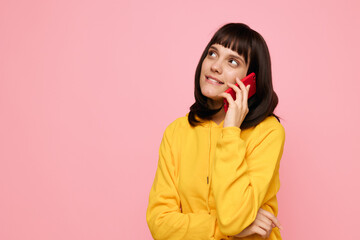 Fototapeta premium A young woman with dark hair and bangs wears a bright yellow hoodie and talks on a red mobile phone against a soft pink background, conveying a cheerful casual mood.