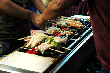 The hands of sellers grill the various types of food such as sauce, mushroom, chilli, pork, ham on the electrical stove in the night flea market