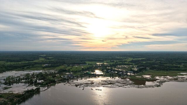 Aerial panoramic view peaceful riverside village in Thailand during sunset featuring calm waters lush green fields and serene sky with soft sunlight reflecting on the river surface.