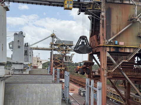 Massive port crane unloads petroleum coke with grab bucket onto conveyor system at modern aluminum production terminal. 444