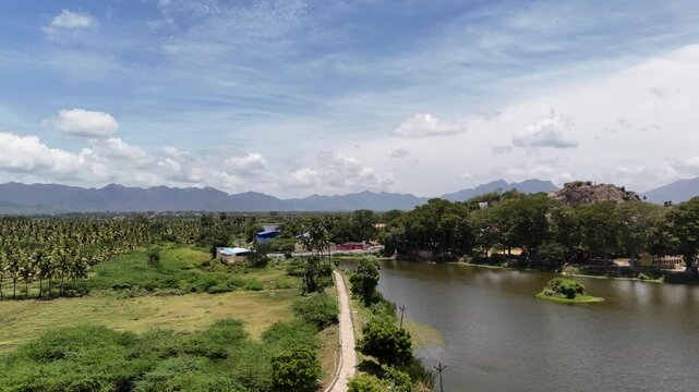 Aerial view of a serene pond surrounded by greenery, palm trees, and distant Western Ghats mountains under a bright blue sky near Tenkasi, Tamil Nadu.