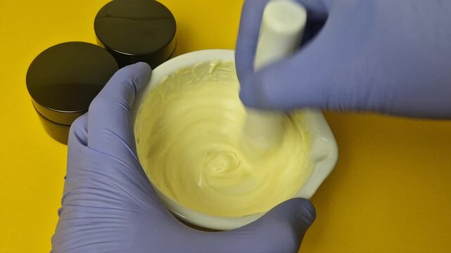 Extreme close-up view of a cosmetic chemist mixing a luxurious, yellow cream base in a white ceramic mortar with a pestle. Hands wear protective blue gloves. Perfect for beauty, pharmacy, and skincare