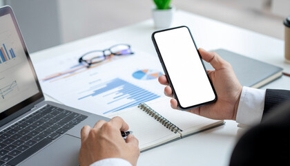 A business person holding a blank smartphone and taking notes over documents with charts and a laptop in a modern office.