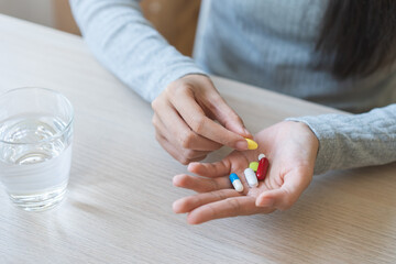 Sick, ill asian young woman, girl hand holding pill capsule, painkiller medicine from stomach pain, headache for treatment, take drug or vitamin with a glass of water at home, pharmacy and health care