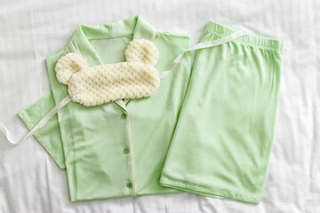 Top view of folded pastel green woman pajama with fluffy sleep eye mask on white bed. Minimal flat lay cozy comfort pyjamas for sleeping and relaxed. Aesthetic lifestyle photo of stylish sleepwear
