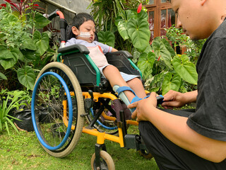 A person helps a child in a wheelchair with leg braces, outdoors in a garden setting, emphasizing...
