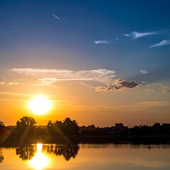 A vibrant sunset casts golden rays over a calm lake, silhouetting trees and distant buildings under a clear sky