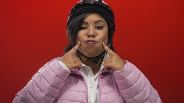 Woman with helmet making a funny face and pointing to her cheeks in front of a red wall wearing a pink jacket.