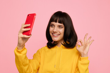 Naklejka premium A young woman in a bright yellow hoodie takes a cheerful selfie against a pink studio background, capturing a confident, vibrant moment of fun and self expression.