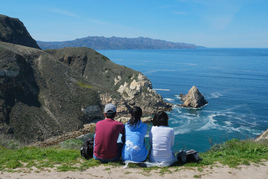 Three people sit on a cliff overlooking the ocean in Channel Islands National Park, California. The rocky coastline, blue water, and distant islands create a peaceful scenic view that captures the bea