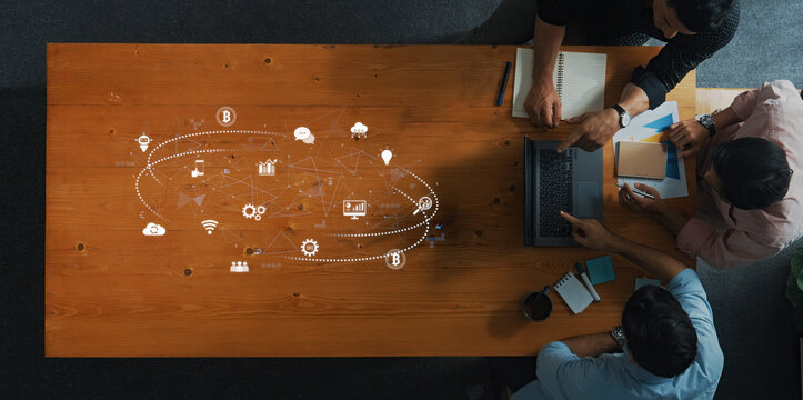 Top view of a collaborative workspace featuring diverse individuals engaged in discussions about digital technology, innovation, and creative ideas on a wooden table. Trope - Powered by Adobe