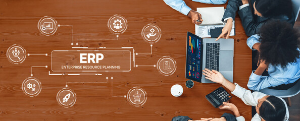 A diverse business team collaborates on an enterprise resource planning project, utilizing laptops and various technology icons on a wooden table. Trope