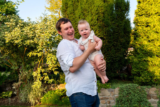 Dad cradles the baby at his side, both laughing softly amidst dense green hedges and stone edging