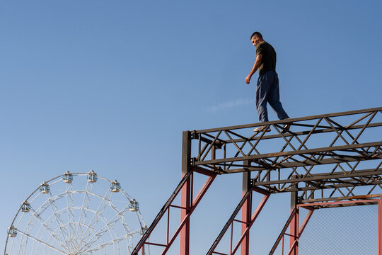 A freerunner walks on a narrow steel beam above the park, with a Ferris wheel in the background. The scene blends urban athleticism with a leisure park environment