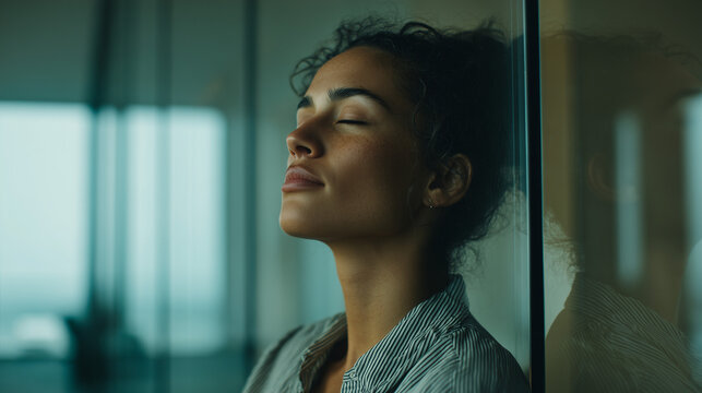 A stressed woman leans against a glass wall with eyes closed, feeling overwhelmed, anxious, and exhausted in a modern office environment, seeking a moment of calm. - Powered by Adobe