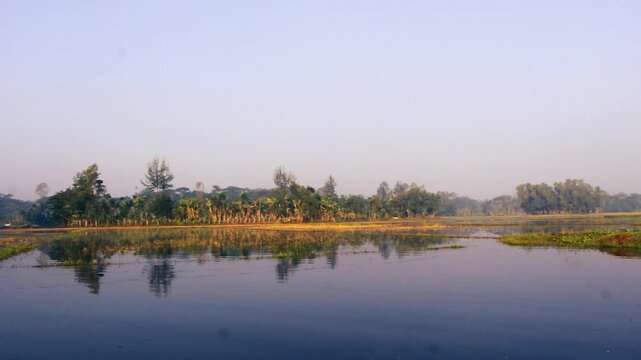 Wide panoramic landscape view of a calm rural river and wetland area during a misty early morning sunrise featuring lush green trees and vegetation on the banks perfectly reflected in the still water 