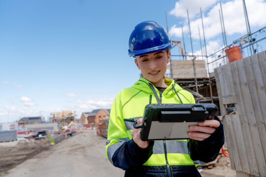 Young woman construction worker in blue helmet checks plans on portable computer tablet at building site