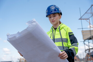 Young woman construction worker in blue helmet checks plans at building site
