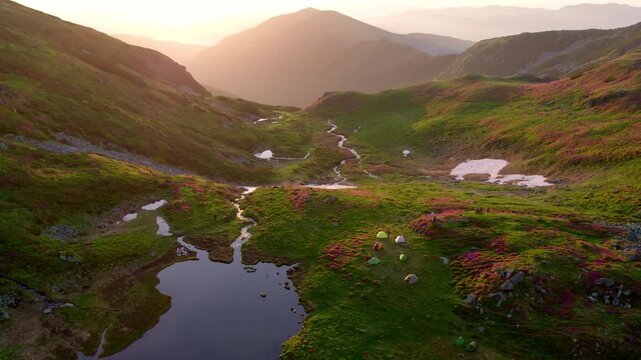Rodnei mountains, Romania. Stunning aerial wide shot of a pristine alpine valley campsite with colorful tents, winding stream, small lake, and patches of snow, illuminated by a warm sunrise.