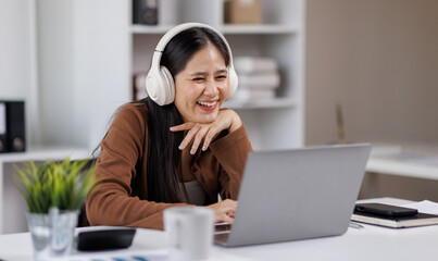 Smiling woman takes notes at her office, wearing headphones listening music at work using laptop...