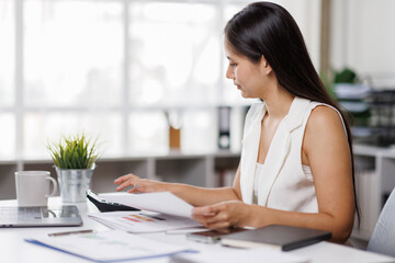 Asian female Using laptop computer and working at office with calculator document on desk, doing planning analyzing the financial report, business plan investment, finance analysis concept.
