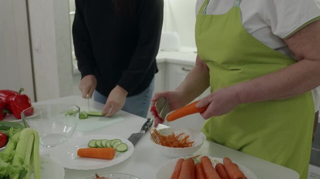 Professional women collaborating in kitchen, grating carrot with peeler and slicing cucumber, demonstrating precise culinary skills and fresh vegetable preparation
