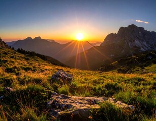 A vibrant sunset over jagged mountain peaks, illuminating a valley filled with grassy fields. The sun casts rays