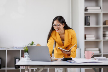 Asian female business woman touching glasses while sitting at workplace near laptop and writing down notes.