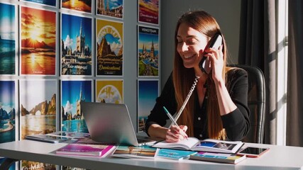 Caucasian travel agent on phone booking vacation packages at bright office desk, smiling while pointing at laptop; travel posters on wall, brochures and notepad on table, sunlight through window