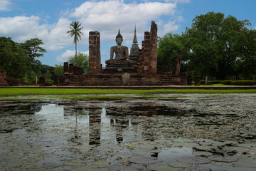 A serene Buddha image stands amid brick ruins beside a lotus-filled pond, creating a tranquil historical sanctuary at Sukhothai Historical Park.