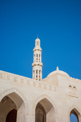 Fototapeta premium A striking view of a white marble mosque featuring a tall minaret and intricately carved arched facade under a bright, cloudless blue sky in Oman. The elegant Islamic architecture and stone carving