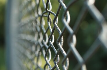 Close up of a weathered chain link fence with a blurred green natural background. The metal has a rough texture showing age and wear. It creates a pattern of diamond shapes fading into the distance.