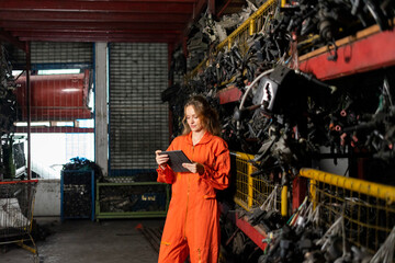 Female technician worker checking inventory with tablet device in garage scrap yard old car part workshop warehouse.