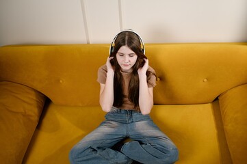 Girl listens to music on headphones while sitting on yellow sofa in relaxed pose, legs crossed, wearing jeans and casual top. Teenager relaxes, listening to calm music, closing her eyes, meditating.