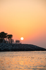 Fototapeta premium A large, bright sun setting or rising over a dark, rocky breakwater, silhouetting palm trees and folded beach umbrellas against a vibrant orange and yellow sky