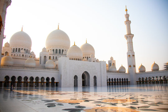 Abu Dhabi, UAE - July 13, 2024: Wide view of the courtyard and exterior of the Sheikh Zayed Grand Mosque, showing the white domes, minaret, and reflective marble floor at sunset
