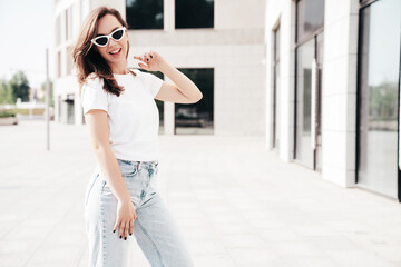 Obraz premium Young beautiful smiling hipster woman in trendy summer white t-shirt and jeans clothes. Sexy carefree model posing on the street background at sunset. Positive model outdoors. In sunglasses