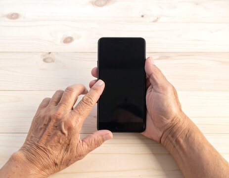 Close-up of elderly hands using a smartphone on a light wood table - Powered by Adobe