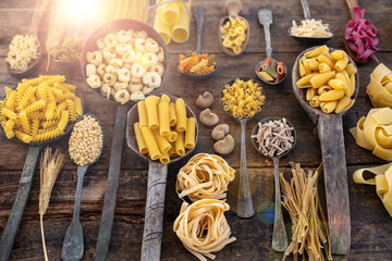 Different types of uncooked pasta on rustic wooden table, in the spoons, cutting board and bowls 