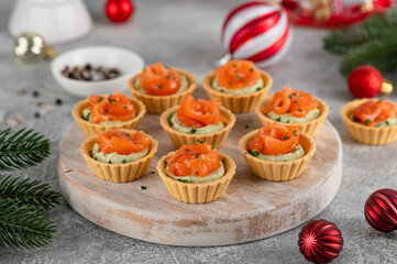 Festive Christmas canapes. Mini tartlets with creamy avocado filling and smoked salmon, garnished with herbs on a rustic wooden board on a gray concrete background. Selective focus. Copy space.