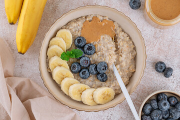 Oatmeal with milk, berries, sliced banana and nut butter in a bowl on a concrete background. Healthy breakfast. Top view.