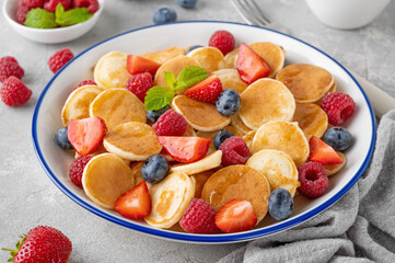 Tiny pancakes with fresh berries, honey and mint for breakfast in a bowl with a cup of coffee on a gray concrete background.