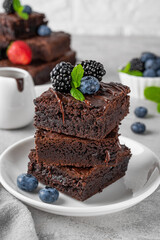 A stack of chocolate brownies with chocolate glaze, fresh berries and mint leaves on top on a white plate on a gray concrete background. Delicious dessert.