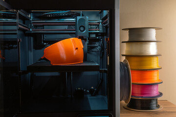 A 3D printer stands on a table with a printed part and a stack of multi-colored plastic next to it.