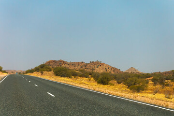 A well maintained road in the Northern Territory of Queensland with a car in the distance, savannah bushland and low hills are to the side.