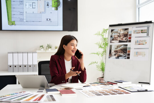 Designer working in office and making call on mobile phone. Female interior designer talking by mobile phone at desk in office. - Powered by Adobe