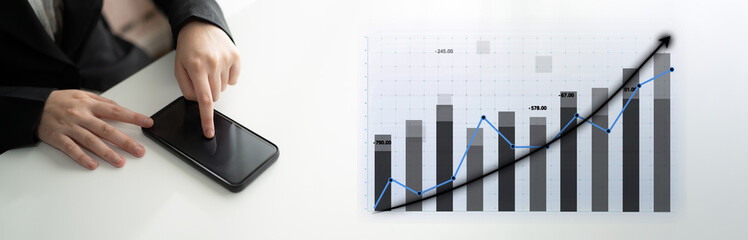 A businessperson interacts with a smartphone while analyzing a graph on a table, representing financial growth and data analysis in a corporate environment. Impute