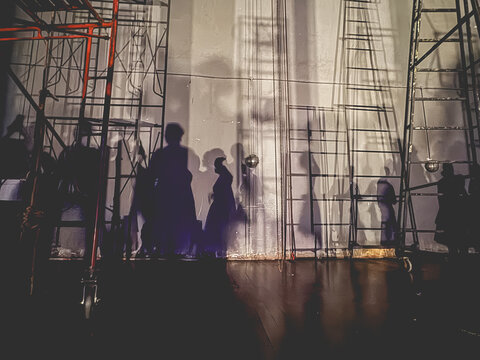 Shadows of ballet dancers and stage crew projected on a backstage wall under dramatic lighting, creating artistic silhouettes and geometric ladder patterns in a theater performance setting.